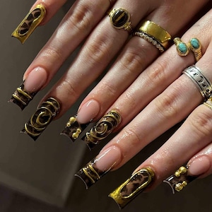 May include: Close-up of a hand with long, sculpted nails featuring a nude base and intricate designs. The nails showcase a combination of gold accents, leopard print, and spiral patterns. Various gold rings with gemstones adorn the fingers.