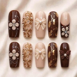 May include: A set of ten press-on nails featuring various designs including dark brown with white floral accents, beige with gold filigree and rhinestones, beige with cream lace bows, dark brown with gold foil, and dark brown with a butterfly.