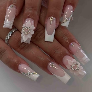 May include: Long, square-shaped acrylic nails with a French manicure design. Some nails feature 3D floral and heart embellishments, while others have rhinestone accents. The nails are painted in a light pink and white color scheme.