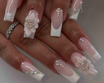 May include: Long, square-shaped acrylic nails with a French manicure design. Some nails feature 3D floral and heart embellishments, while others have rhinestone accents. The nails are painted in a light pink and white color scheme.