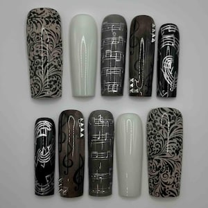 May include: A set of ten artificial nails in various designs. Some nails feature musical notes in silver, others have floral patterns in black and gray, and some are solid white or gray.