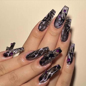 May include: Long, coffin-shaped acrylic nails with a dark, smoky design. The nails feature black chain accents, purple gems, and a spider charm on one nail. The overall design is dark and gothic.