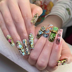 May include: A hand displaying long, ornate, and colorful acrylic nails. Each nail features a unique design with gold accents, gemstones, and intricate patterns. The color palette includes pink, green, blue, and gold, creating a luxurious and artistic aesthetic.