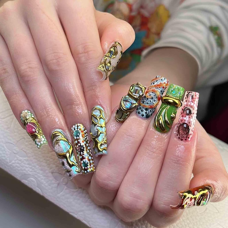 May include: A hand displaying long, ornate, and colorful acrylic nails. Each nail features a unique design with gold accents, gemstones, and intricate patterns. The color palette includes pink, green, blue, and gold, creating a luxurious and artistic aesthetic.