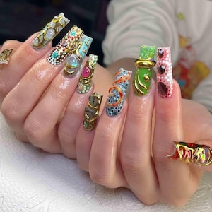 May include: A hand with long, ornate nail art. The nails feature a variety of designs, including gold embellishments, gemstones, and intricate patterns in colors like white, blue, green, and red. The nails are a statement of style and creativity.