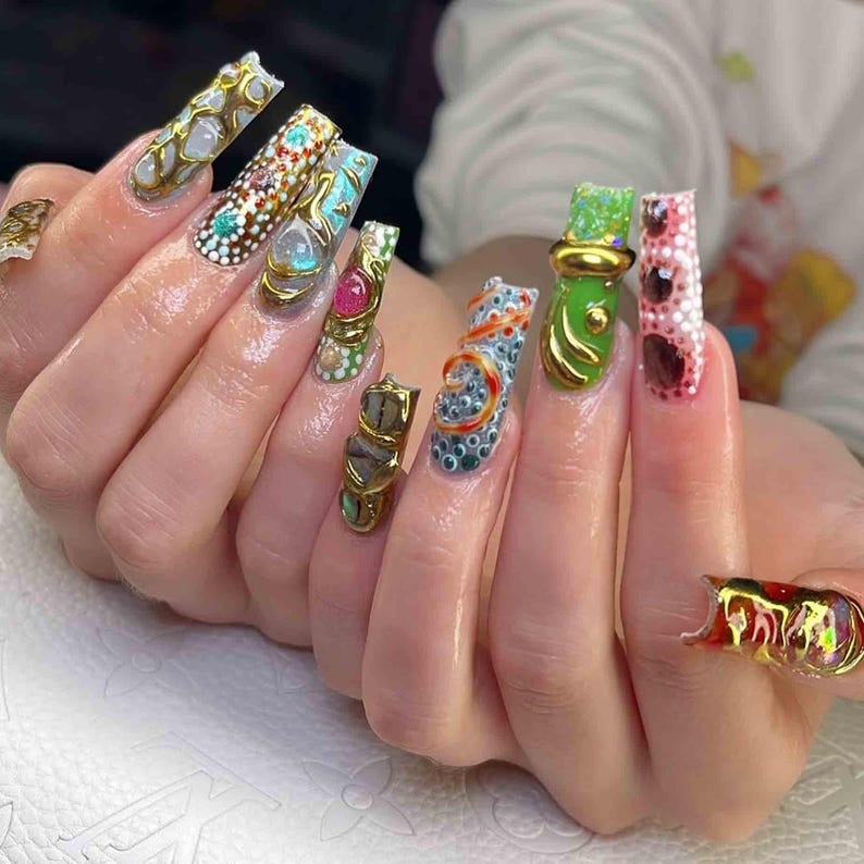 May include: A hand with long, ornate nail art. The nails feature a variety of designs, including gold embellishments, gemstones, and intricate patterns in colors like white, blue, green, and red. The nails are a statement of style and creativity.