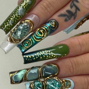 May include: Close-up of long, decorated fingernails. The nails feature various designs, including gold embellishments, glitter, and shades of green and blue. Some nails have a swirl design, while others have a gemstone-like appearance.