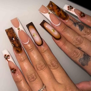 May include: Long, square-shaped acrylic nails in various designs. Some nails feature a tortoiseshell pattern, others have a white base with a nude oval, and some have a brown gradient. Gold embellishments and flower accents add detail.