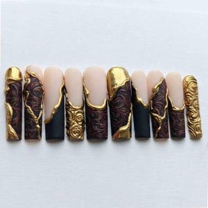 May include: A set of ten press-on nails featuring a mix of matte black and nude bases. The designs include intricate raised gold filigree patterns, smooth gold metallic accents, and deep red embossed floral swirls across the various nail tips.