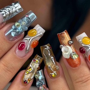 May include: Close-up of a set of long, embellished acrylic nails. The nails feature a variety of designs, including silver metallic accents, colorful gems, and textured gel effects. The color palette includes clear, orange, yellow, red, and silver.