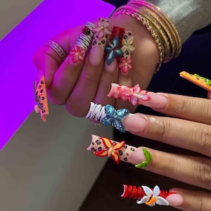 May include: A hand displaying long, decorated acrylic nails. The nails feature various designs, including floral accents, animal prints, and vibrant colors like red, blue, and yellow. Some nails have 3D flower embellishments.