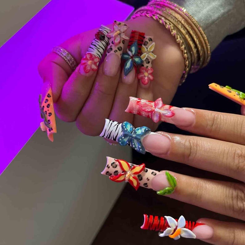 May include: A hand displaying long, decorated acrylic nails. The nails feature various designs, including floral accents, animal prints, and vibrant colors like red, blue, and yellow. Some nails have 3D flower embellishments.