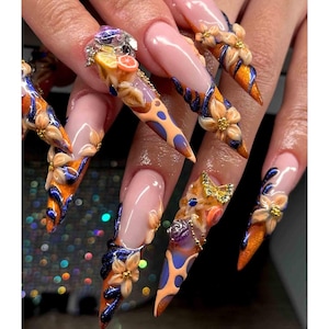 May include: Long, pointed artificial nails with a nude base and intricate designs. The nails feature orange and blue accents, floral embellishments, and some have fruit and gem details. The overall effect is ornate and artistic.