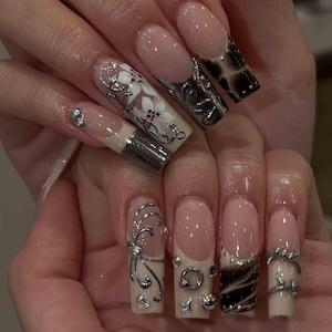 May include: Long square-shaped artificial nails featuring a nude base with French tips. The designs include white floral patterns, metallic silver swirls, chrome-like black and silver gradients, small silver studs, and a silver ring piercing.