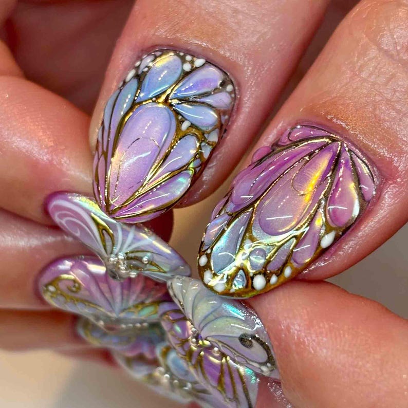 May include: Close-up of a hand with intricately designed nail art. The nails feature a butterfly wing design with iridescent purple, blue, and gold accents. The design is detailed with gold outlines and white dots, creating a 3D effect.