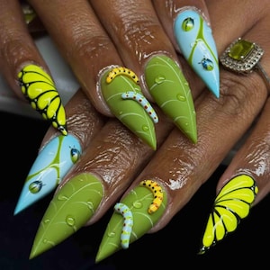 May include: A set of long, pointed fingernails with a nature-inspired design. The nails feature a variety of designs, including green leaf patterns with 3D caterpillars, butterfly wings, and blue and green accents. The nails are painted with bright colors and have a glossy finish.