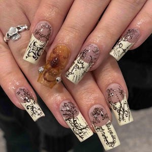 May include: Acrylic nails with a distinctive design. The nails are long and square, featuring clear, beige, and brown hues, with detailed black line art and embellishments. One nail has a 3D sculpted design with rhinestones, and some nails have the word "Angel".
