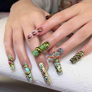 May include: A close-up of a hand with long, elaborately decorated fingernails. The nails feature various designs, including animal prints, gemstones, and metallic accents. The color palette includes green, gold, red, and blue.