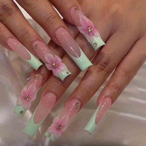 May include: Long, square-shaped artificial nails with a light pink base and a French tip in a soft mint green. Each nail features a 3D pink flower with gold accents and small rhinestones.