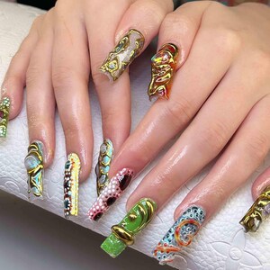 May include: A close-up of a set of long, sculpted acrylic nails. Each nail features a unique design with gold accents, including textured surfaces, colorful patterns, and abstract shapes. The nails are a mix of colors, including gold, green, red, and blue.