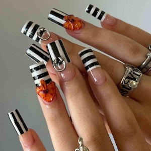 May include: Long, square-shaped artificial nails with a clear base and black and white striped design. Some nails feature silver rings and orange flower accents. The nails are adorned with a glossy top coat.