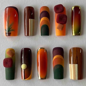 May include: A collection of ten artificial nails with various designs. The nails feature a range of colors, including orange, brown, green, and gold. Some nails have geometric patterns, while others have textured elements or miniature plant illustrations. The nails are rectangular in shape.