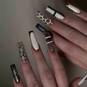May include: A set of long, artificial nails with various designs. Some nails are white with gold trim, others feature a black and gold design, and one has a 3D bubble pattern. The nails showcase a modern, artistic aesthetic.