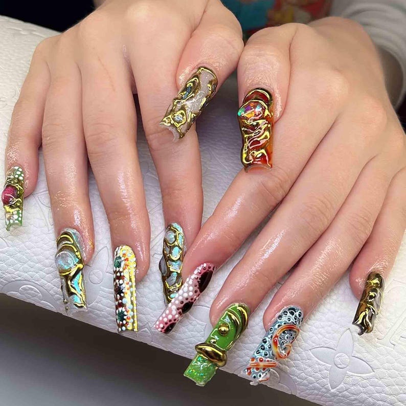 May include: A close-up of a hand with long, elaborately decorated fingernails. Each nail features a unique design with gold, green, red, and blue accents. The nails have a glossy finish and are intricately detailed.