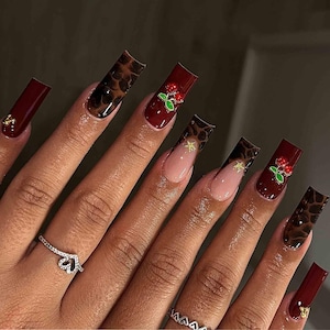 May include: A hand with long, square-shaped acrylic nails. The nails feature a variety of designs, including a dark red solid color, a leopard print, and a French tip with gold accents. Some nails have cherry embellishments.
