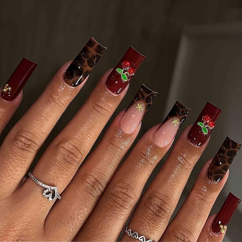 May include: A hand with long, square-shaped acrylic nails. The nails feature a variety of designs, including a dark red solid color, a leopard print, and a French tip with gold accents. Some nails have cherry embellishments.