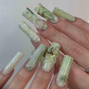 May include: A set of long, light green and white acrylic nails. Some nails feature floral designs, while others have 3D embellishments of leaves and pearls. The nails are well-manicured and have a glossy finish.