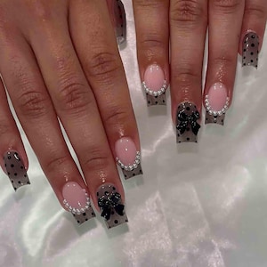May include: A set of manicured nails featuring a light pink oval shape with a pearl border. The nails have a gray polka dot design with black bow accents. The nails are square shaped.