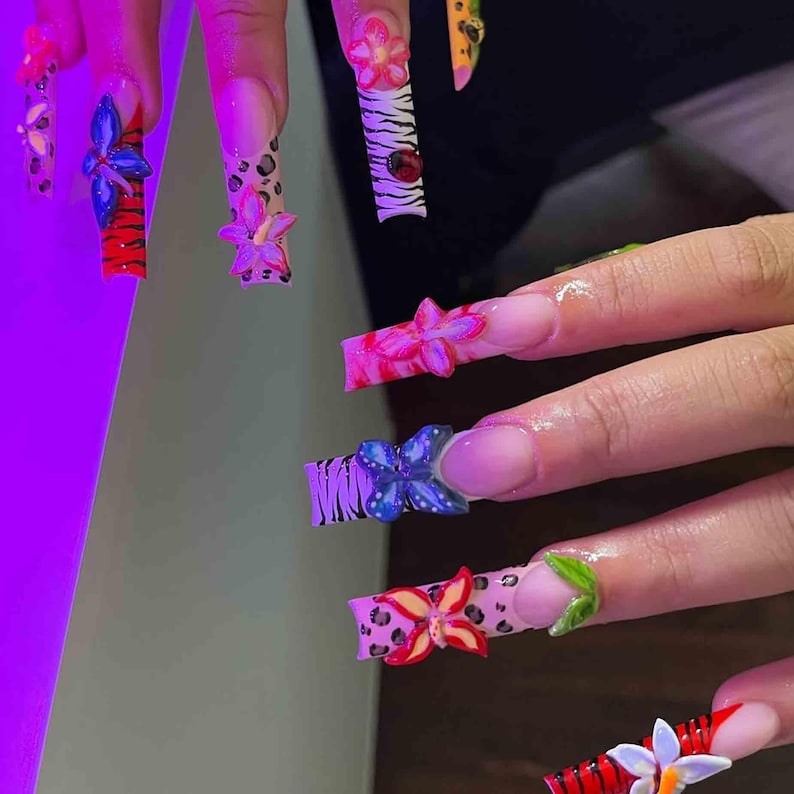 May include: A set of long, decorated fingernails. Each nail features a different design, including floral embellishments, animal prints, and vibrant colors like red, pink, blue, and yellow. The nails are a form of artistic expression.