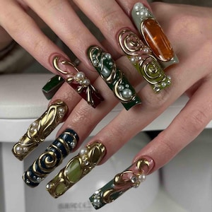 May include: A set of long, ornate artificial nails. Each nail features a unique design with gold accents, pearls, and various colors including green, burgundy, and navy. Some nails have a swirl design, while others have a more linear pattern.