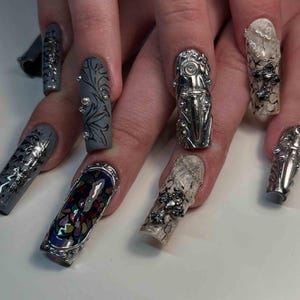 May include: A close-up of a hand with long, embellished fingernails. The nails feature various designs, including silver metal accents, floral patterns, and colorful mosaic-like designs. The nails are painted in shades of gray, clear, and multi-colored.
