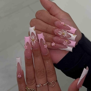 May include: Long, acrylic nails with a pink and white design. The nails feature a French tip with a pink wave design, white accents, and small gold embellishments. Some nails have 3D flower accents.