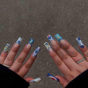 May include: Long square-shaped artificial nails featuring a white base with intricate blue and red floral patterns. Some nails include gold-toned evil eye charms and small metallic accents. The design is consistent across all ten fingernails.