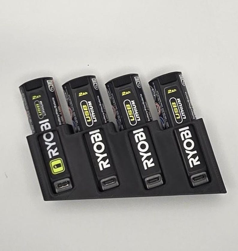 May include: A black 3D-printed battery holder with four Ryobi USB Lithium batteries. Each battery is black with white and yellow text, including "2Ah" and "USB Lithium". The holder has a slot for each battery.
