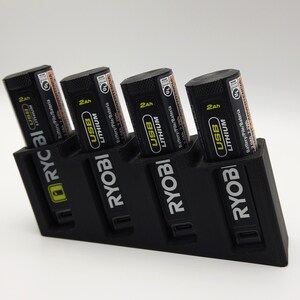 May include: A black battery charging station holding four black and orange USB Lithium batteries. The batteries are labelled with "2Ah" and "RYOBI" branding. The charging station also has the "RYOBI" logo.