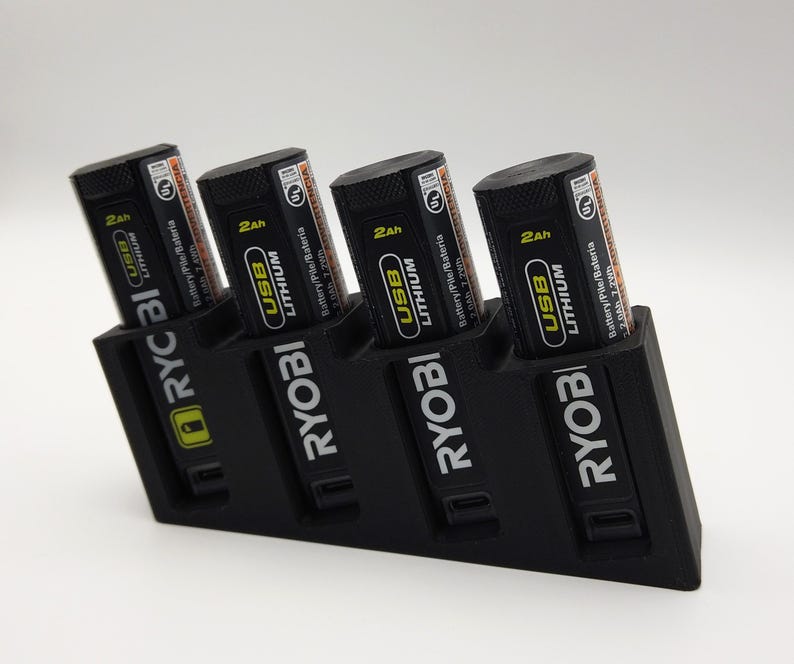 May include: A black battery charging station holding four black and orange USB Lithium batteries. The batteries are labelled with "2Ah" and "RYOBI" branding. The charging station also has the "RYOBI" logo.