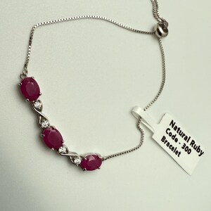 May include: A silver chain bracelet with a sliding clasp. The bracelet features four oval-shaped ruby gemstones with smaller clear gemstones on either side. The tag attached to the bracelet reads "Natural Ruby Code - 300 Bracelet".