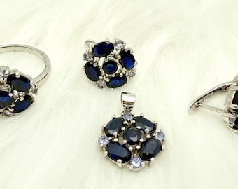 May include: A jewelry set featuring a ring, pendant, earrings, and a cufflink. Each piece has a silver setting and is adorned with dark blue oval-cut gemstones and smaller white gemstones, arranged in a floral pattern.