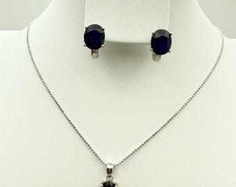 May include: A jewelry set featuring a silver necklace with a dark blue oval pendant, matching earrings, and a ring. The earrings and ring have dark blue oval stones. The set is displayed on a white stand.