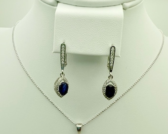 May include: A silver jewelry set featuring a ring, earrings, and a necklace. Each piece has a dark blue, oval-shaped stone surrounded by smaller clear stones. The ring and earrings have a similar design, while the necklace has a pendant.