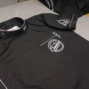 Peut inclure: Chemise noire à manches courtes avec un col blanc et une bordure blanche. La chemise a une poche sur le côté gauche avec un logo brodé blanc qui dit "Barbershop Cousins" avec une paire de ciseaux et le texte "Haircuts & Shaves".