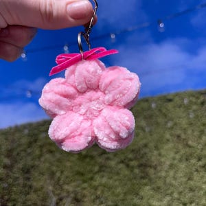 Puede incluir: Un llavero de flor rosa peluda con un lazo rosa. El llavero está unido a un anillo de metal plateado.