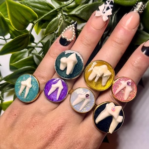 Puede incluir: Una colección de siete anillos circulares con dijes blancos en forma de diente sobre fondos de resina coloridos y brillantes en tonos verde azulado, verde oscuro, morado, amarillo, azul claro, rosa y negro, con algunos anillos con cristales.