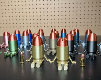 May include: A collection of small, anthropomorphic bullet-shaped figurines with metallic copper-colored tips and bodies in various colors including green, gold, black, blue, and silver. Each figurine has two small arms and legs. Two real brass cartridges stand in front.