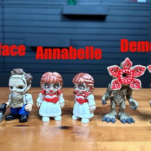Puede incluir: Seis peque&ntilde;as figuras coleccionables alineadas. Dos figuras de Leatherface con motosierras, dos mu&ntilde;ecas Annabelle con vestidos blancos y detalles rojos, y dos figuras de Demogorgon con cabezas rojas en forma de flor. El texto Leatherface, Annabelle y Demogorgon aparece en rojo sobre las figuras.