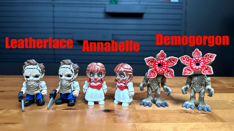 Puede incluir: Seis peque&ntilde;as figuras coleccionables alineadas. Dos figuras de Leatherface con motosierras, dos mu&ntilde;ecas Annabelle con vestidos blancos y detalles rojos, y dos figuras de Demogorgon con cabezas rojas en forma de flor. El texto Leatherface, Annabelle y Demogorgon aparece en rojo sobre las figuras.